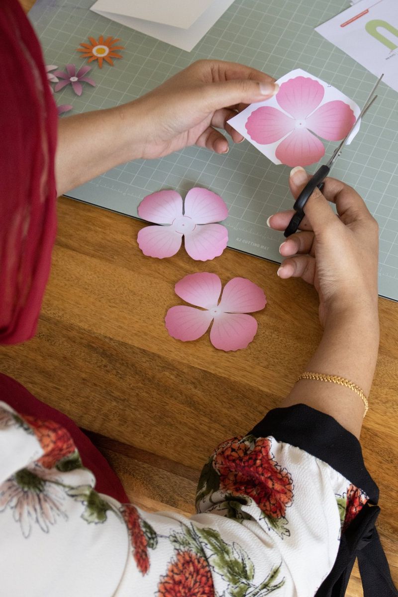 Les mains d'une femme découpant des fleurs en papier roses avec des ciseaux.