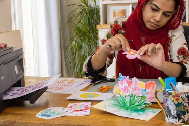 Une femme tient délicatement une fleur en papier sur une table jonchée de cartes colorées faites à la main et d'outils de fabrication, et sur laquelle une imprimante est posée.