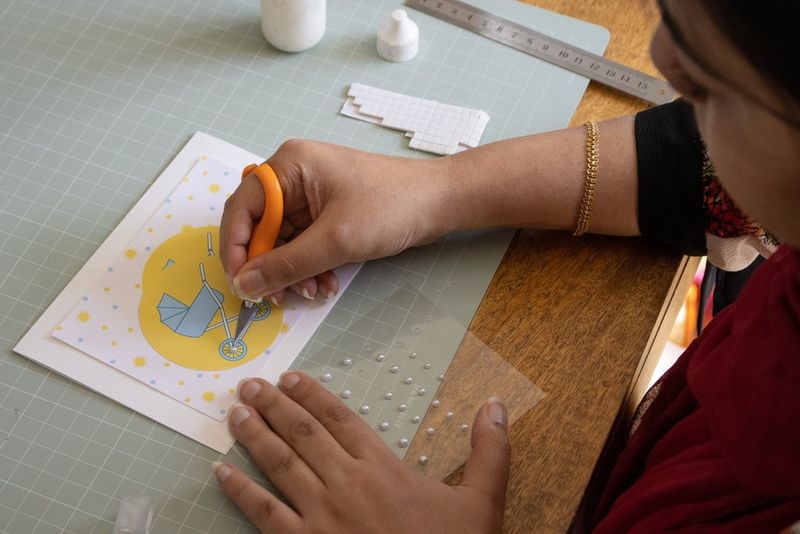 Une femme ajoutant des perles argentées aux roues d'une poussette sur une carte de vœux faite à la main.