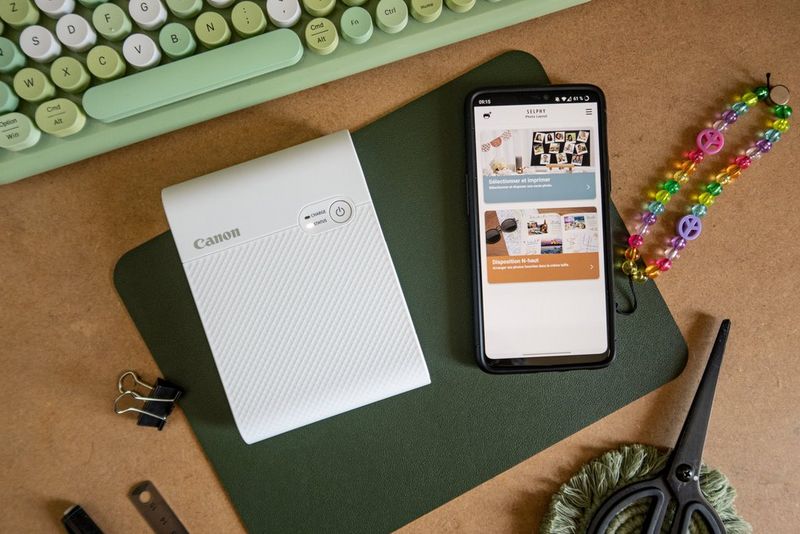 Een telefoon met de Canon SELPHY Photo Layout-app op een bureau naast een draagbare Canon SELPHY Square QX10-printer en een telefoonbedel met kralen.