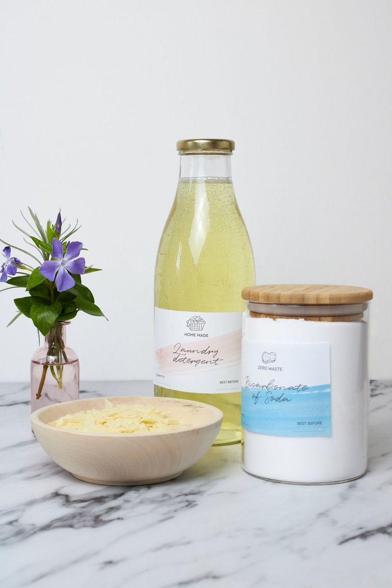A bottle of detergent and a jar of bicarbonate of soda, both with handmade labels, on a marble worktop next to a vase of flowers and a bowl of grated cheese.
