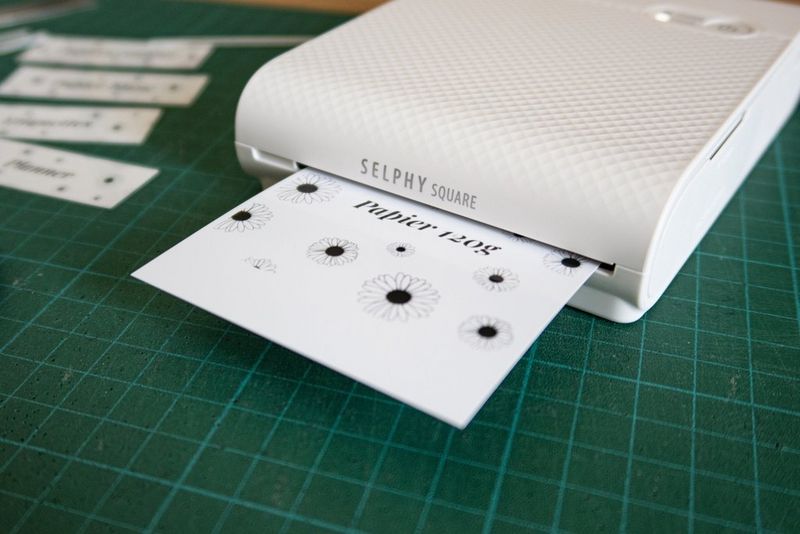 A printed homemade label with sunflower motifs and text in a cursive font emerging from a Canon SELPHY Square QX10 portable printer.