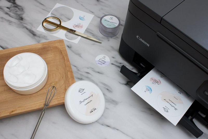 A sheet of homemade labels emerging from a printer, next to some scissors and cut-out labels, and the lid of a cosmetics container with a printed label.