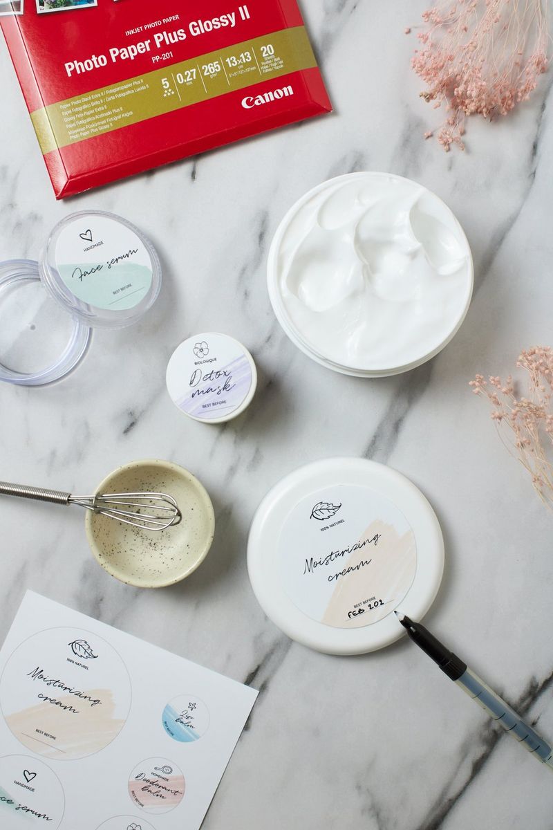 A pack of printer paper on a worktop alongside a sheet of printed homemade labels and several small cosmetics containers with printed labels.