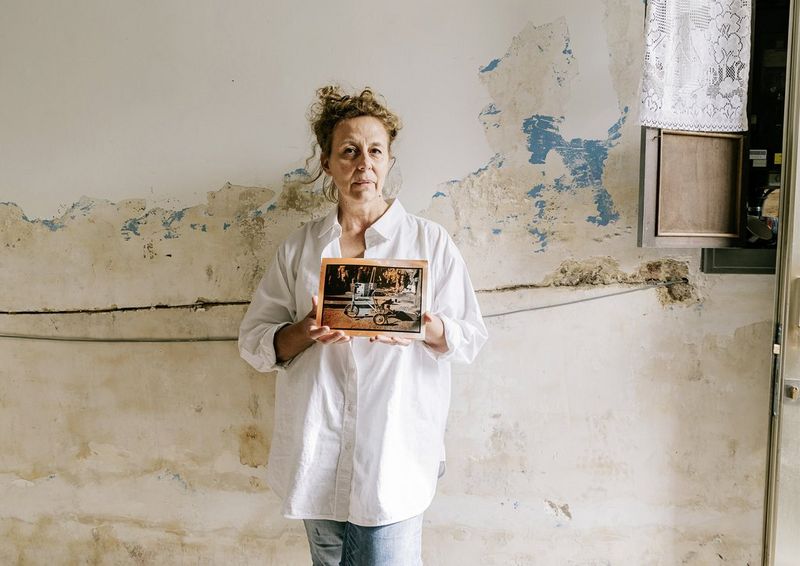 אישה עומדת מול קיר ומחזיקה תמונה מוכתמת של תלת-אופן בשחור-לבן.