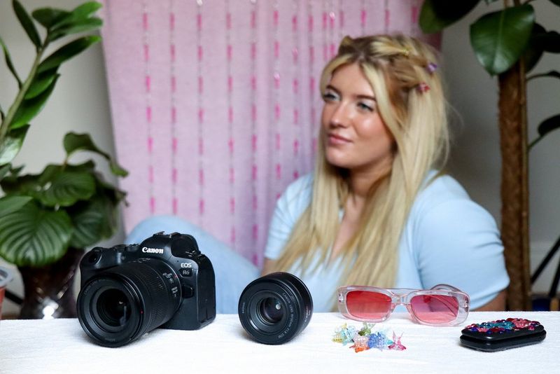 Ženska z dolgimi svetlimi lasmi sedi pred rožnatim blagom, na katerem visijo nizi kroglic. Pred njo so Canonov fotoaparat in objektiv, zaprt telefon na preklop, ki je prekrit z nalepljenimi dragulji, rožnato obarvana sončna očala in sponke v obliki metulja.