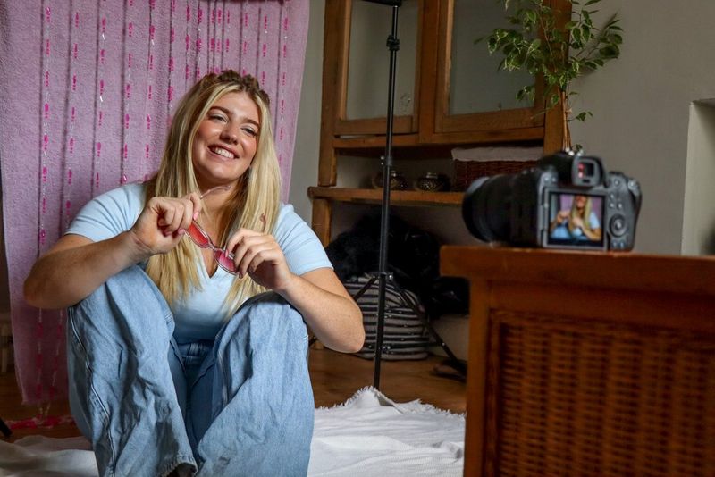 Ženska z dolgimi svetlimi lasmi sedi na tleh dnevne sobe, drži rožnato obarvana sončna očala in se smehlja proti fotoaparatu, ki je nameščen na leseni mizi.