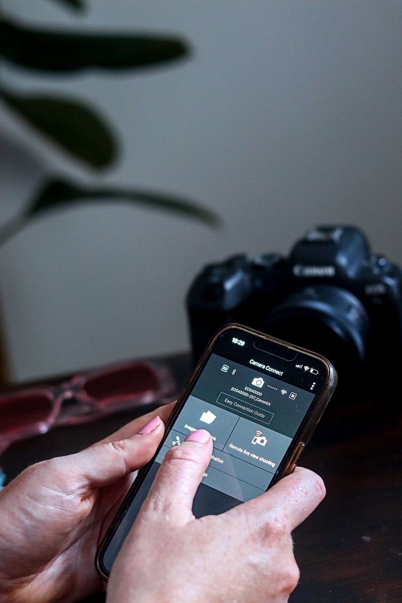 Roke držijo pametni telefon, ki prikazuje program Canon Camera Connect, s Canonovim fotoaparatom na mizi zadaj.