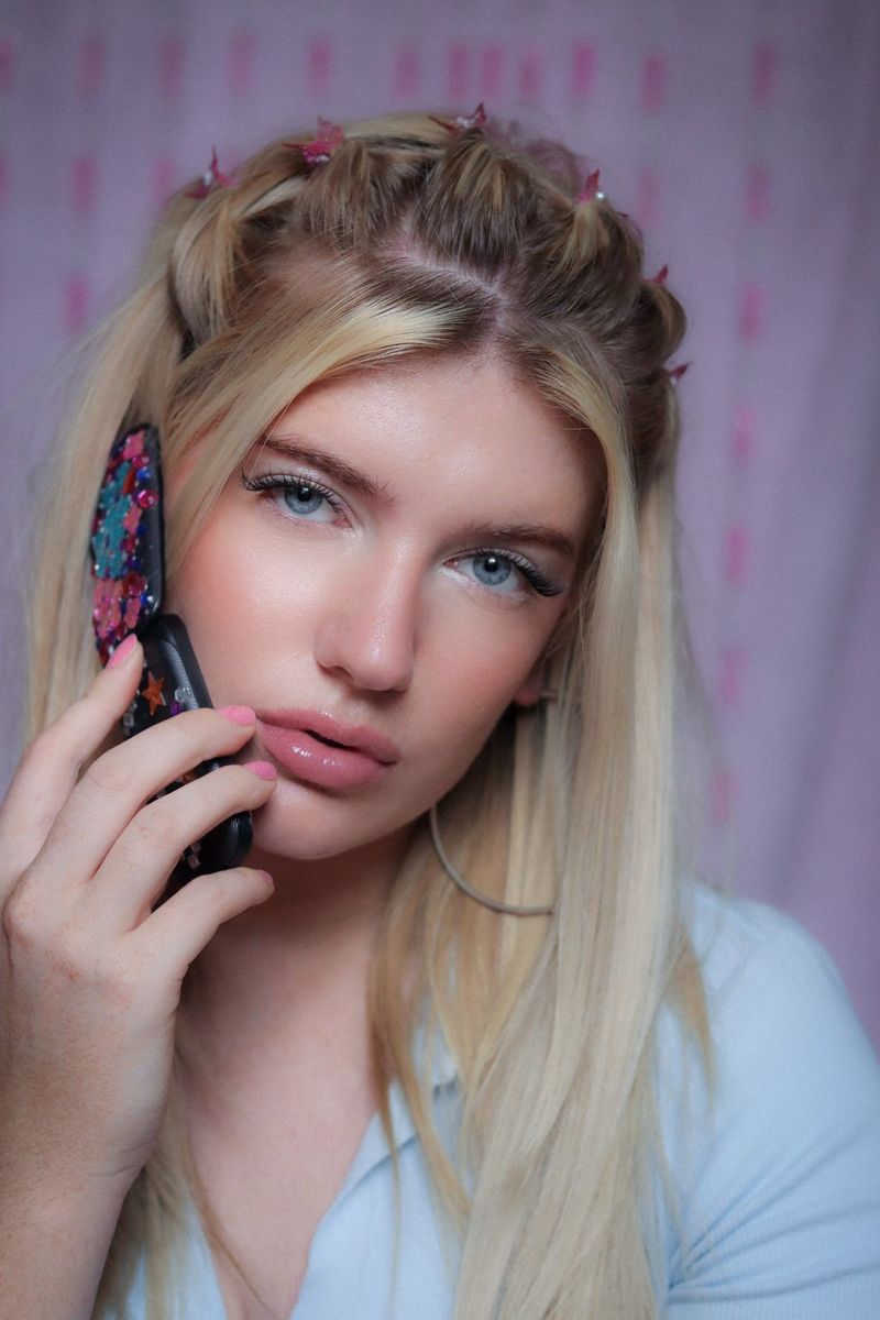 Ženska z dolgimi svetlimi lasmi, ki so na vrhu speti z več majhnimi sponkami v obliki metulja, drži telefon na preklop ob strani obraza in se mršči v fotoaparat.