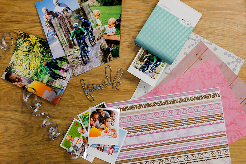 Принтер SELPHY Square QX10 печатает изображения семьи на велосипедах. На столе разложены листы бумаги с разноцветными узорами и фотографии той же семьи в разных форматах.