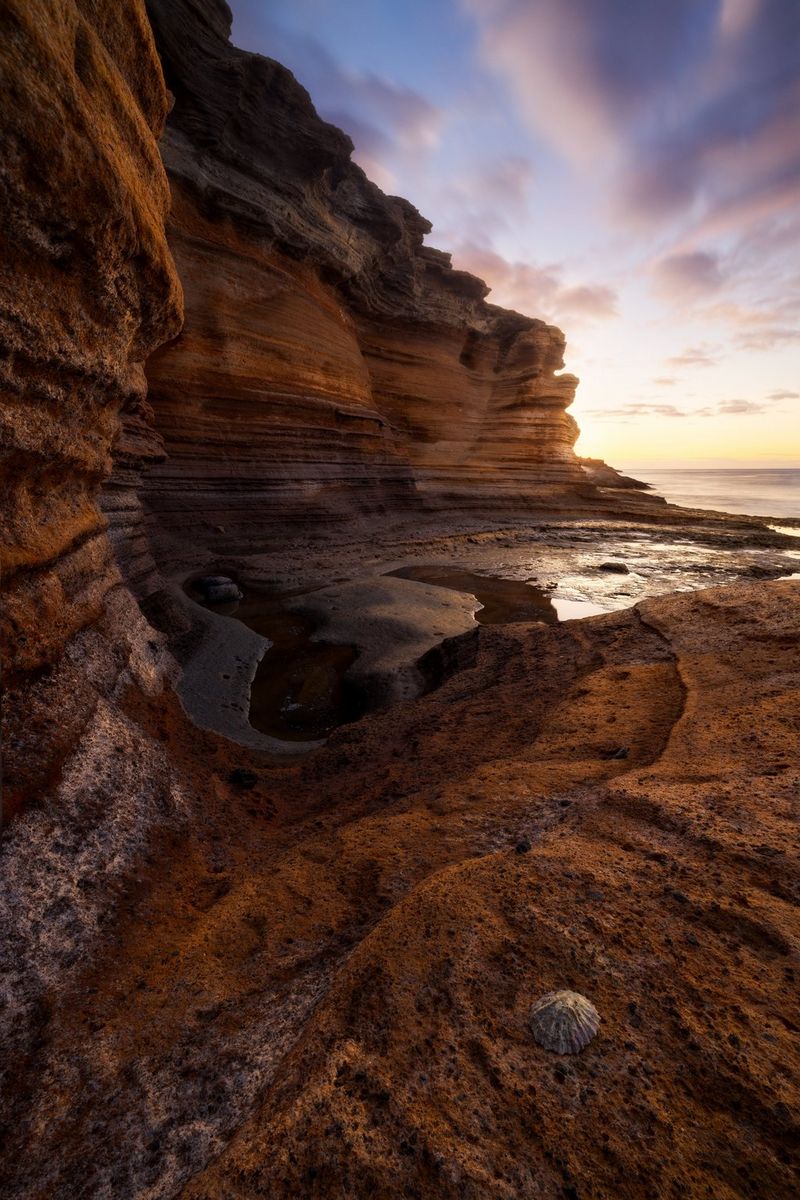 Сделанная Эриком Коломбо фотография обрывистого утеса изогнутой формы и ракушки на камнях на переднем плане — все это на фоне слоистых облаков.
