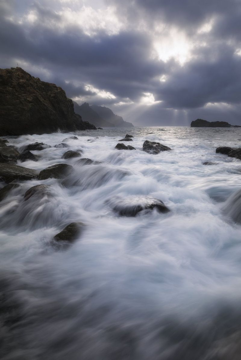 На этой составной фотографии HDR, созданной Эриком Коломбо, размытые с помощью длительной выдержки волны накатывают на скалы. Темно-серые облака на небе закрывают солнце.