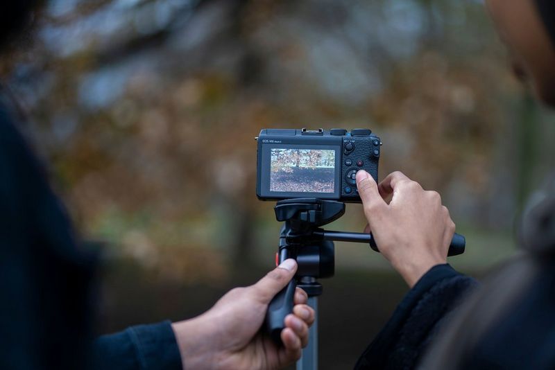 Două mâini ajustând setările pe partea din spate a unui aparat foto Canon EOS M6 Mark II montat pe trepied.