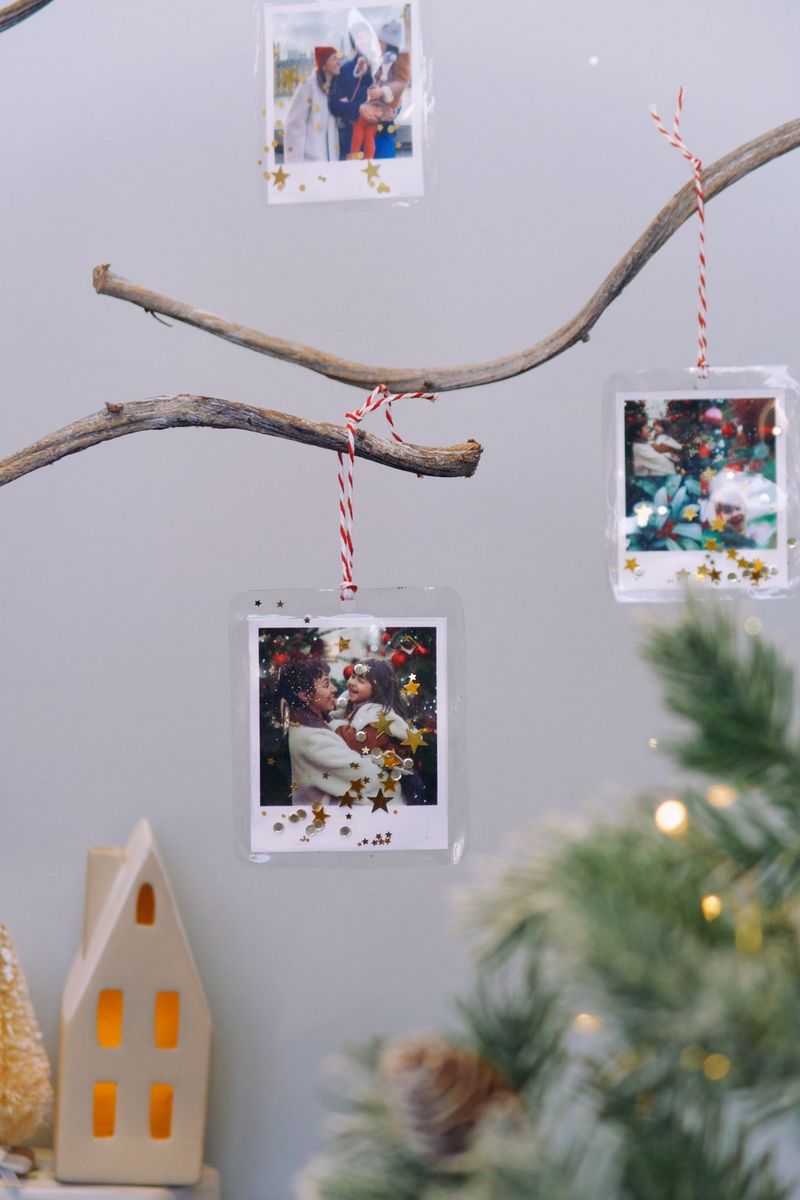 Три новогодних украшения из фотографий, украшенных блестками и распечатанных на принтере Canon SELPHY Square QX10, подвешены на ветках новогодней елки рядом с бумажной фигуркой домика.