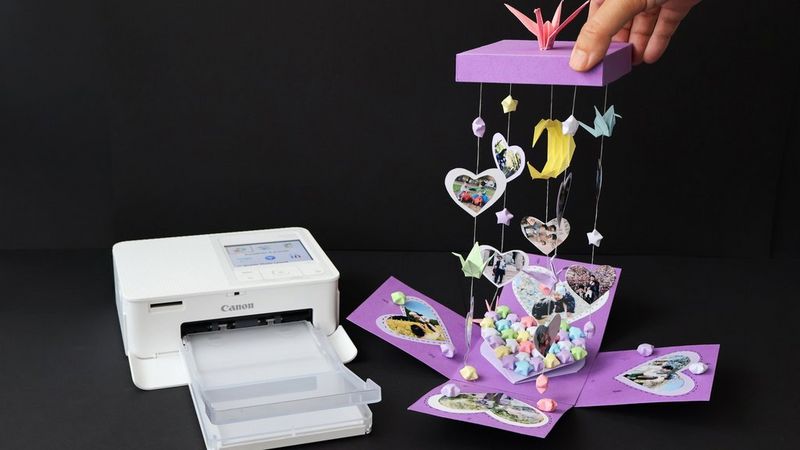 Una stampante Canon SELPHY si trova accanto a una scatola a sorpresa viola riempita di stelle di carta, decorata con fotografie a forma di cuore e con altre fotografie e origami appesi al coperchio.