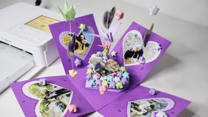Una scatola a sorpresa viola riempita di stelle di carta, decorata con fotografie a forma di cuore e con altre fotografie e origami appesi al coperchio. Accanto c'è una stampante Canon SELPHY.