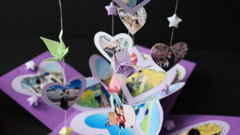 Una scatola a sorpresa viola riempita di stelle di carta, decorata con fotografie a forma di cuore e con altre fotografie e origami appesi al coperchio.