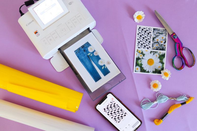 Margherite di stoffa, un foglio con immagini di margherite e la foto di un paio di jeans con margherite su una superficie viola, accanto a una stampante Canon SELPHY CP1300.