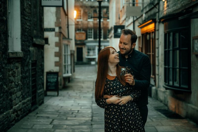 Мужчина стоит позади женщины и обнимает ее — они стоят посреди аллеи со множеством исторических зданий.