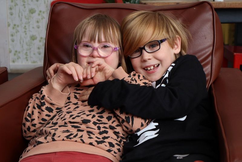 בתמונה שצולמה באמצעות Canon EOS R100, שני ילדים חייכנים מתחבקים בשובבות כשהם יושבים יחד על כורסה.