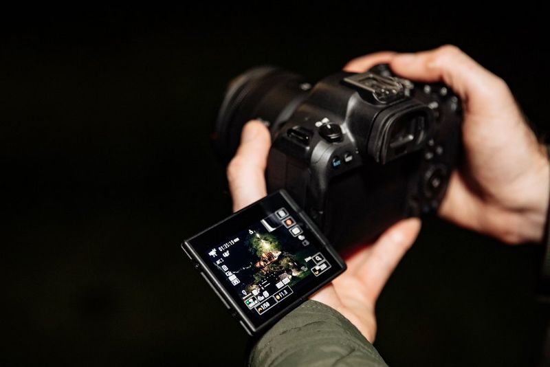 Par ruku drži EOS R6 Mark II tvrtke Canon čiji je dodirni zaslon promjenjivog kuta nagiba rasklopljen prema van i u stranu te prikazuje snimku drvene kolibe u šumi.