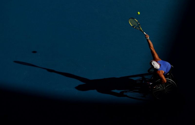 Eine Person im Rollstuhl greift nach oben, um einen Tennisball zu schlagen, wobei sich ihr Schatten vor einem blauen Hintergrund nach links erstreckt.