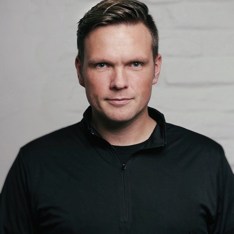 Headshot of a person in a black top in front of a white brick background.