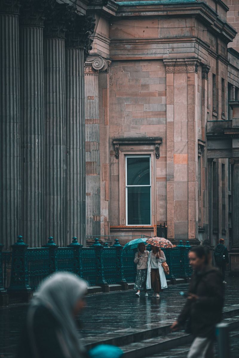 Street photographer Andres McNeill captures a street photography image of two figures holding umbrellas walking in front of a large, imposing building.