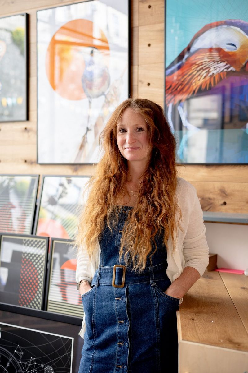 Een vrouw met rood haar in een denimjurk staat voor schilderijen aan een muur met houten wandbetimmering.