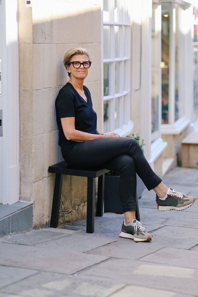 Een vrouw met kort blond haar, een bril en donkere kleding zit op een kruk buiten een lichtgekleurd stenen gebouw.