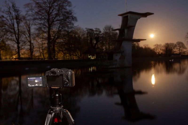 A Canon EOS RP camera set up on a tripod to photograph the Moon over a lake. An old diving board rises high above the water.