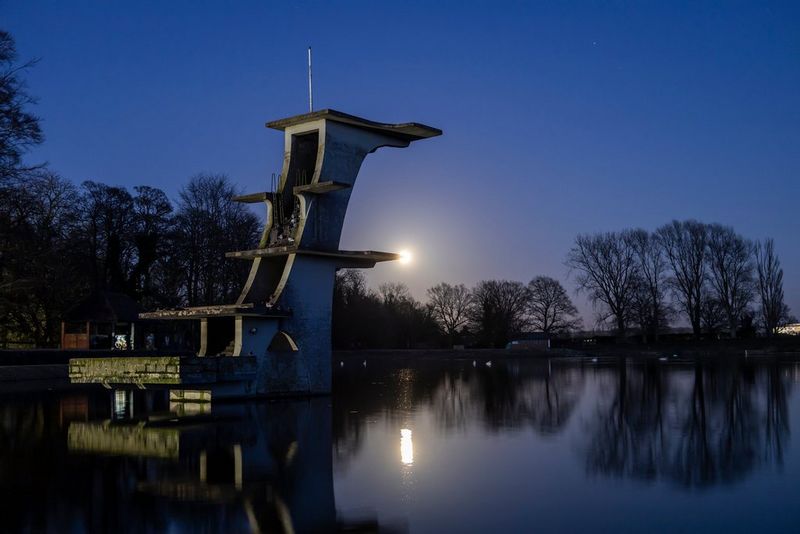 The Moon reflecting out over a lake. An old diving board rises high above the water.
