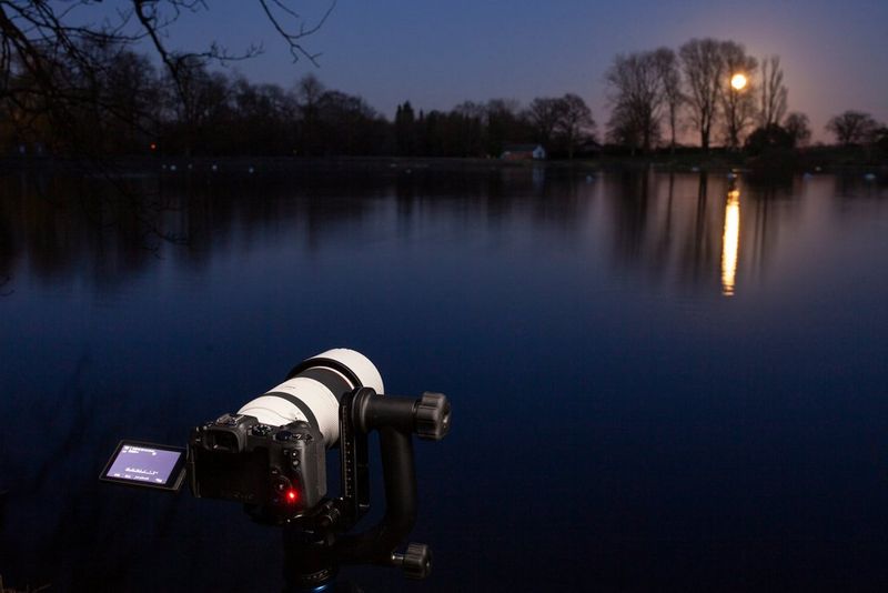 An EOS RP on a tripod with a telephoto lens, set up to photograph the Moon rising above the tree-line around a lake.