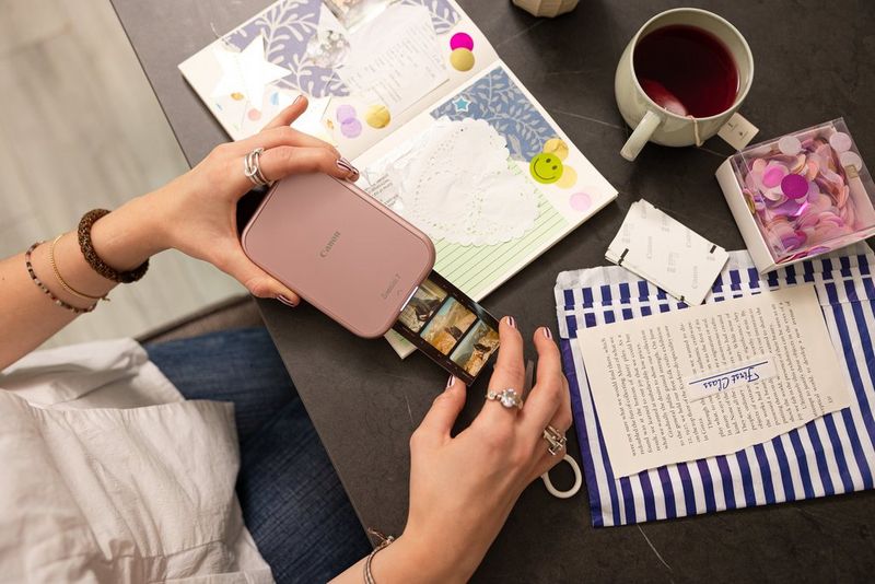 Elde tutulan Canon Zoemini 2 yazıcıdan bir fotoğraf şeridi çıkıyor; masanın üzerinde ise bir karma günlük ve çeşitli diğer kağıt ürünleri duruyor.
