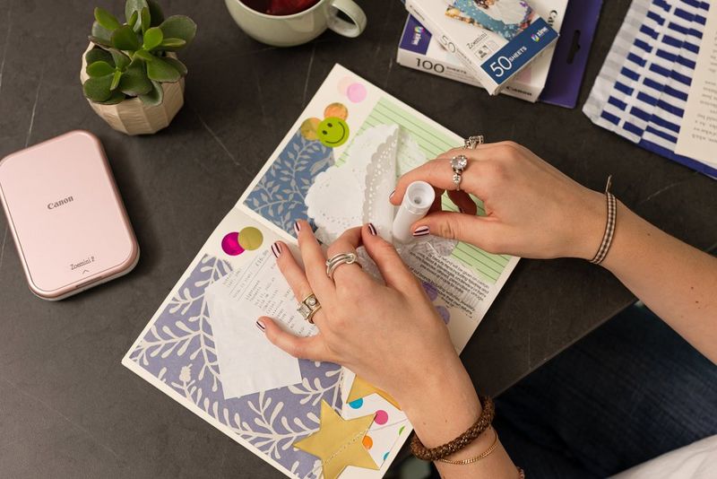 Genç bir kadının elleri, kalp şeklinde bir kağıt peçeteyi bir karma günlüğe yapıştırıyor. Yandaki masada ise Canon Zoemini 2 taşınabilir yazıcı duruyor.