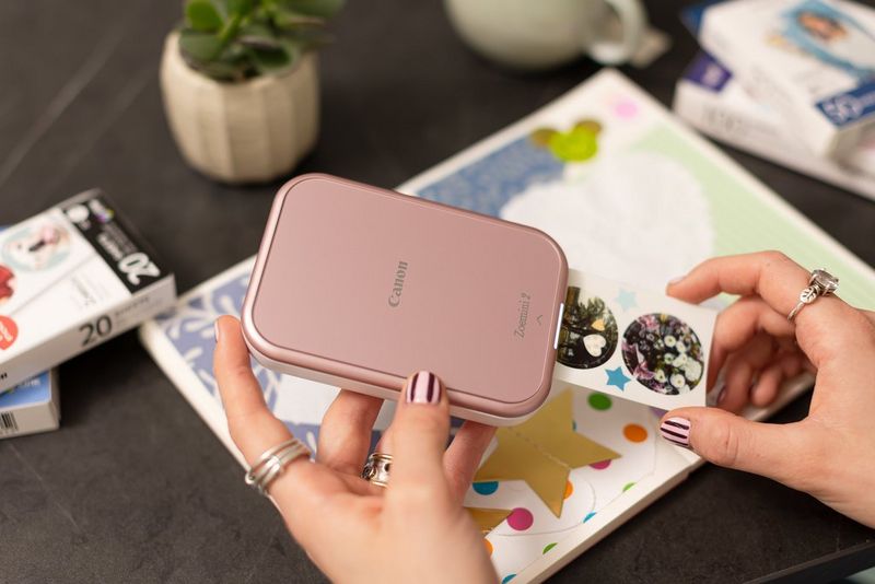 Kullanıcının elinde tuttuğu Canon Zoemini 2 taşınabilir yazıcıdan iki yuvarlak çıkartmaya basılmış görüntülerin bulunduğu bir sayfa çıkıyor, masanın üzerinde ise bir karma günlük duruyor.