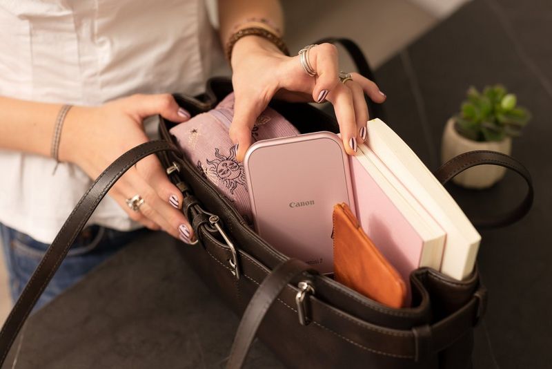 Bir kullanıcı, kompakt Canon Zoemini 2 yazıcıyı iki küçük fotoğraf albümü ve diğer eşyalarla bir el çantasına koyuyor.