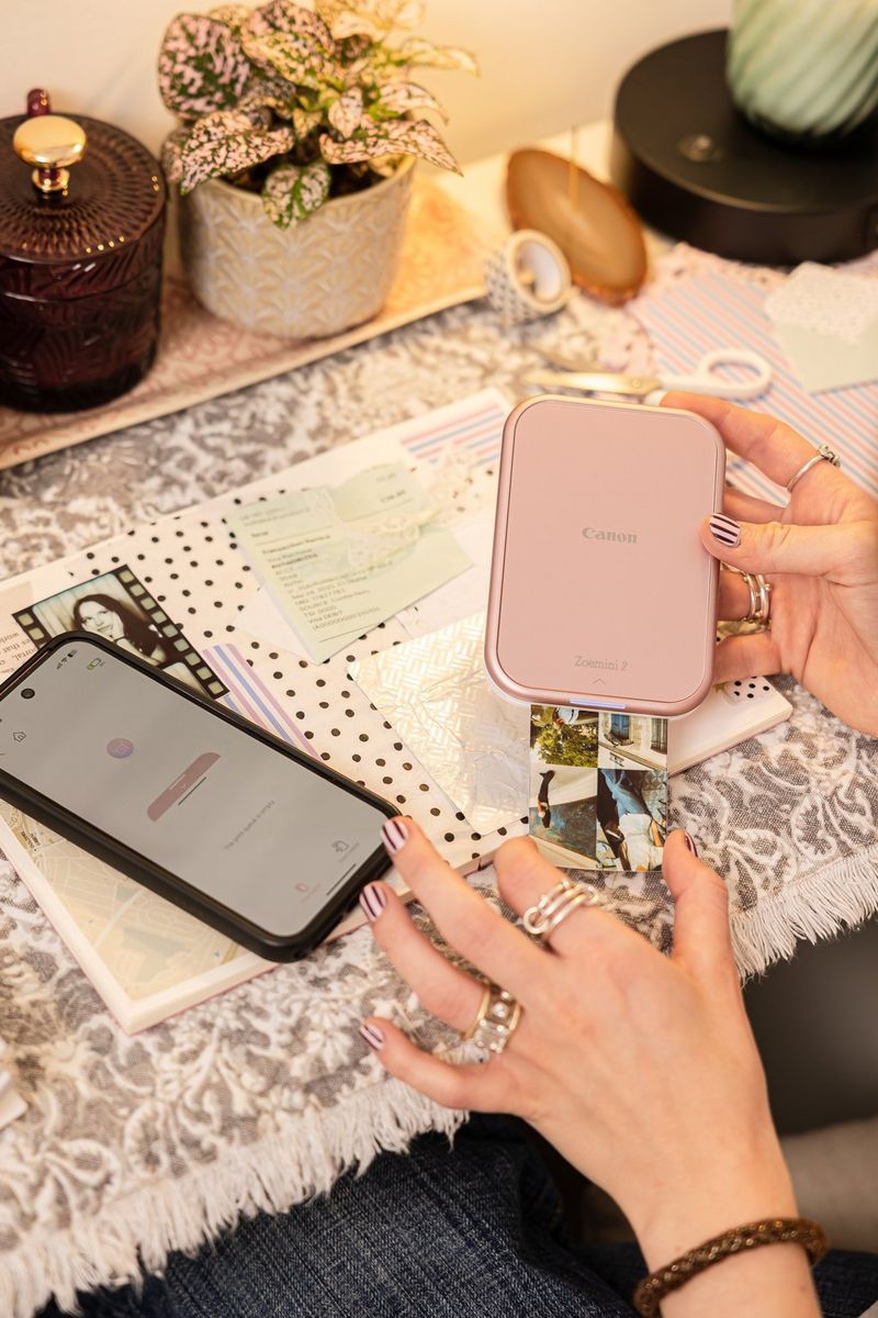 Kullanıcının elinde tuttuğu Canon Zoemini 2 yazıcıdan küçük fotoğrafların kolajını içeren bir baskı çıkıyor. Masanın üzerinde ise Canon Mini Print uygulamasının açık olduğu bir akıllı telefon duruyor.