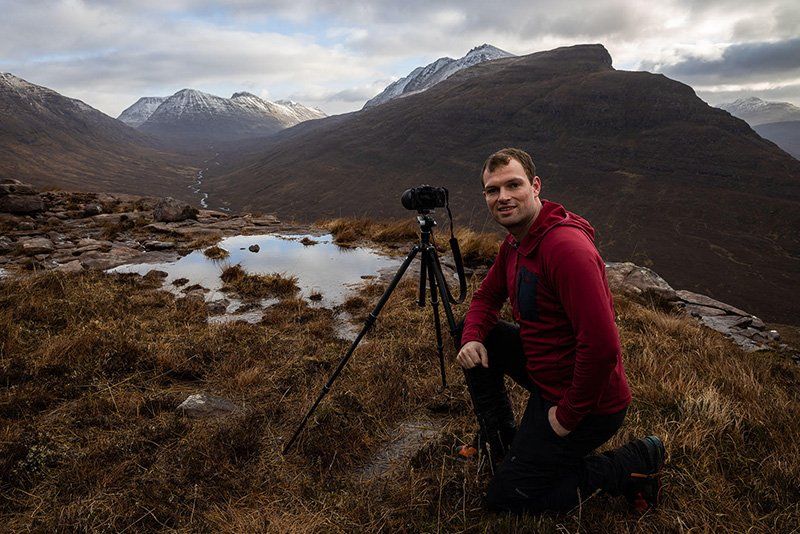 Snow blows through the air creating a mist, covering a mountain in front of a river.
