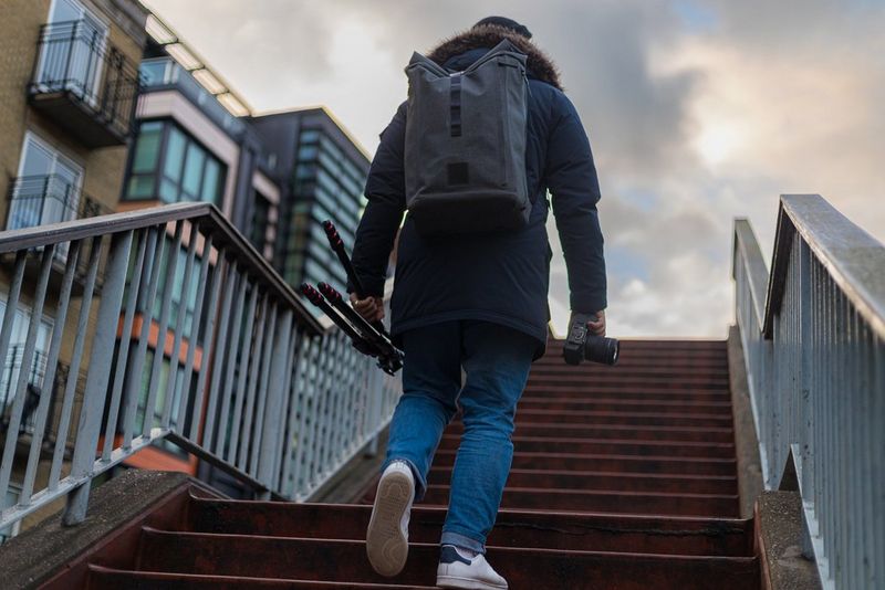 Muž, který má na sobě kabát a batoh, stoupá po schodech venku ve městě a v jedné ruce drží fotoaparát Canon EOS R6 a v druhé stativ.