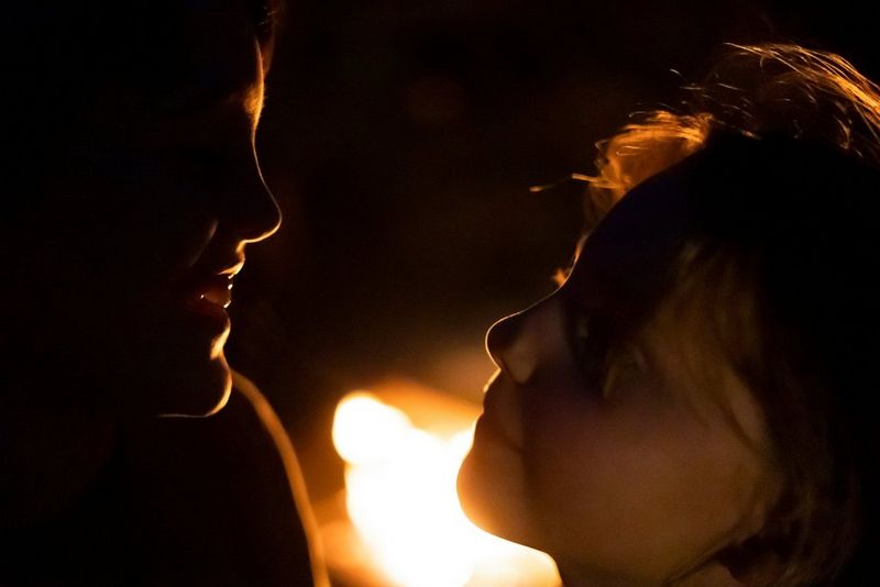 שני ילדים פנים אל פנים מול מדורה. המתאר של הצדודיות שלהם מואר על-ידי האור שמגיע מהלהבות.