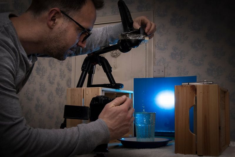 Un homme tient son appareil photo au-dessus d'une tasse remplie d'eau installée contre une feuille bleue pendant qu'il photographie une goutte qui tombe.