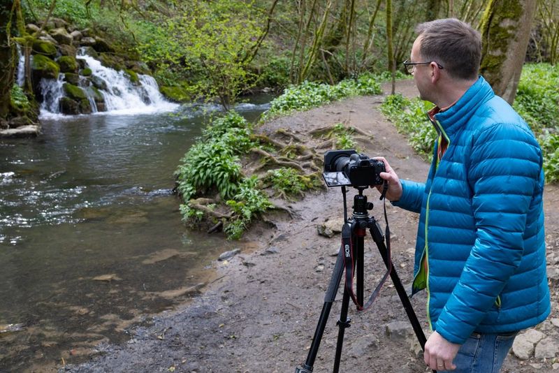 Un photographe se tenant devant une rivière avec une chute d'eau, son appareil photo sur un trépied.