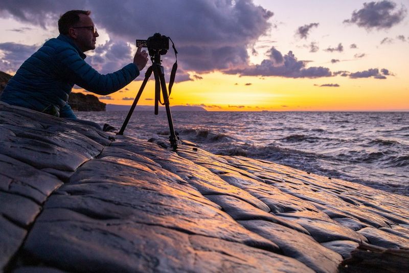 Un homme accroupi à côté d'un appareil photo et d'un trépied sur une plage couverte de rochers.