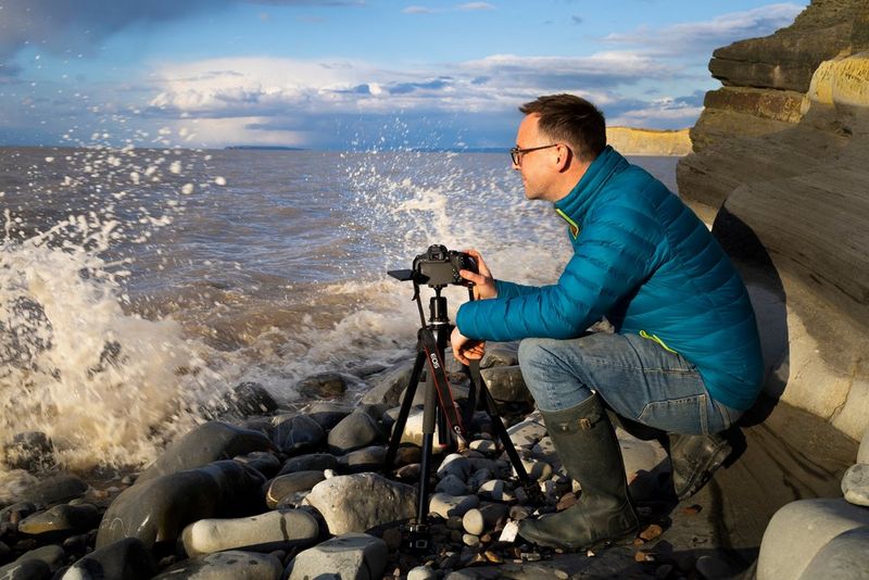 Un photographe accroupi sur les rochers happés par les vagues avec son trépied et son appareil photo.