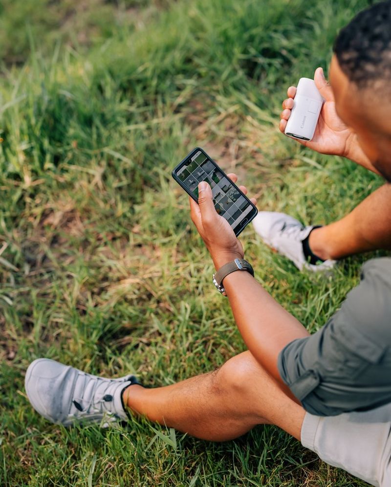 A man sat down outdoors, looking at photos of wildlife on his phone, holding a Canon PowerShot ZOOM