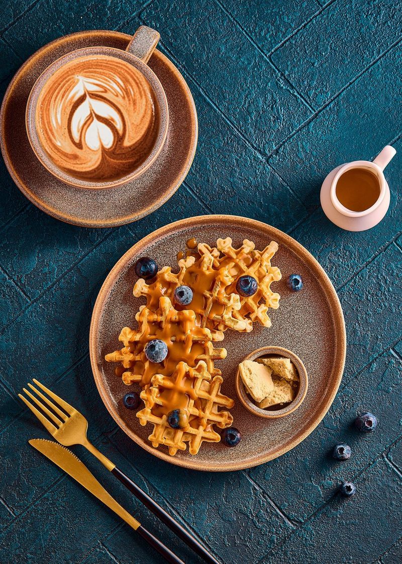 Bovenaanzicht van wafels, gegarneerd met blauwe bessen en karamel, geserveerd naast een kop koffie, kan, mes en vork, gepresenteerd op een grijze leisteen.
