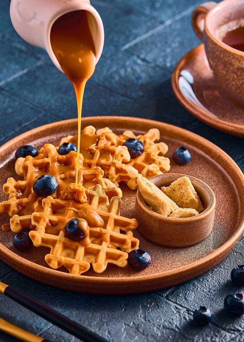 Een kan giet dikke karamel over wafels die zijn gegarneerd met blauwe bessen en liggen op een grijze leisteen.