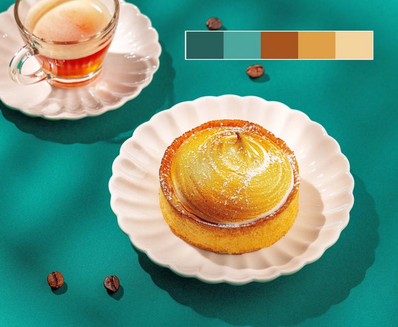 Een wit geschulpt bord met een kleine citroenmeringuetaart naast een kop thee tegen een groene achtergrond met koffiebonen en een kleurenkaart in de rechterbovenhoek.