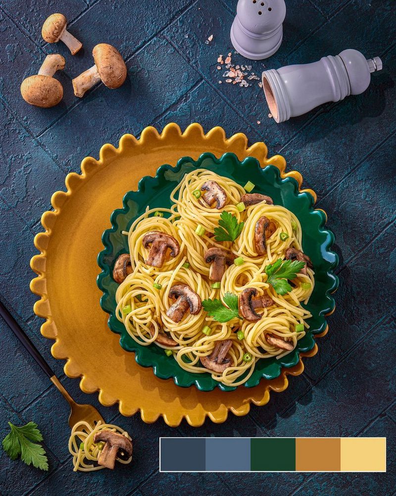 Een bord met spaghetti-nestjes met stukjes champignons en groene blaadjes op een donkergroene plaat met een kleurenkaart in de rechterbenedenhoek.